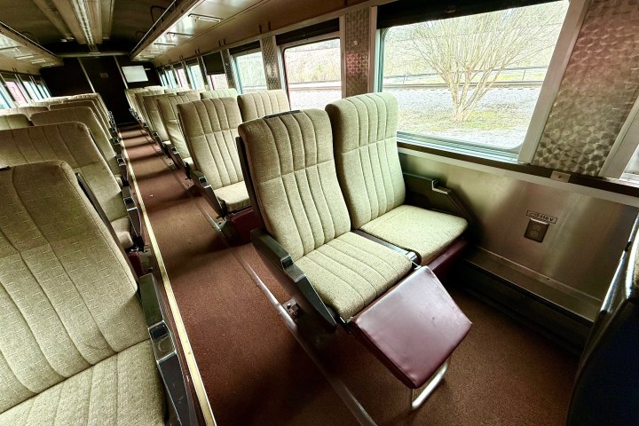 Interior of an empty train carriage with rows of beige upholstered seats and large windows.