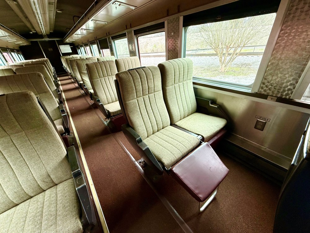 Interior of an empty train carriage with rows of beige upholstered seats and large windows.