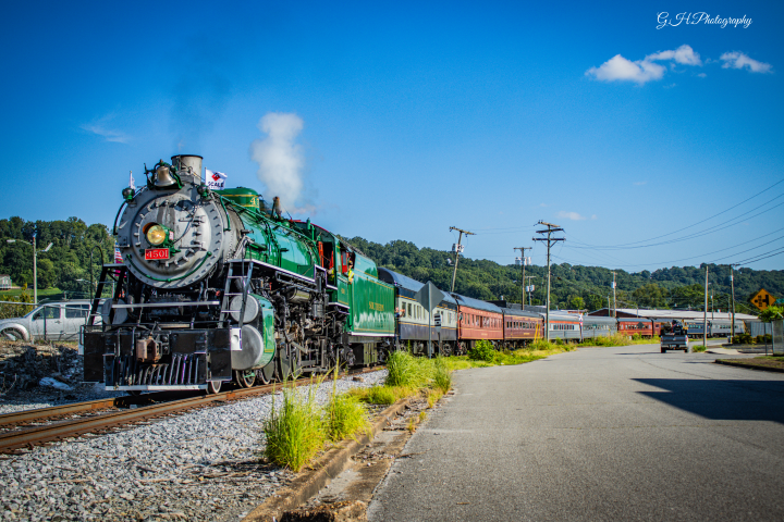 Vintage green steam train on track near road, blue sky above.