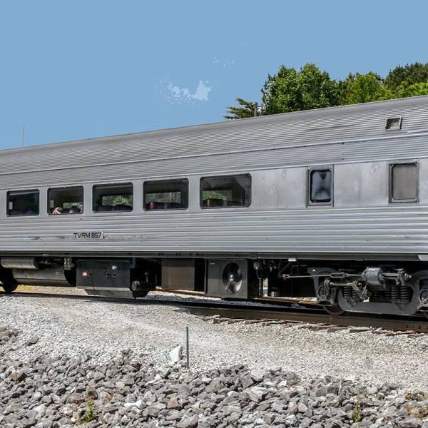 Stainless steel passenger railcar labeled TVRM 857 on a sunny day, parked on gravel near a wooded area.