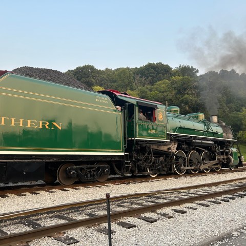 Vintage steam locomotive on tracks, emitting smoke, with trees in the background.