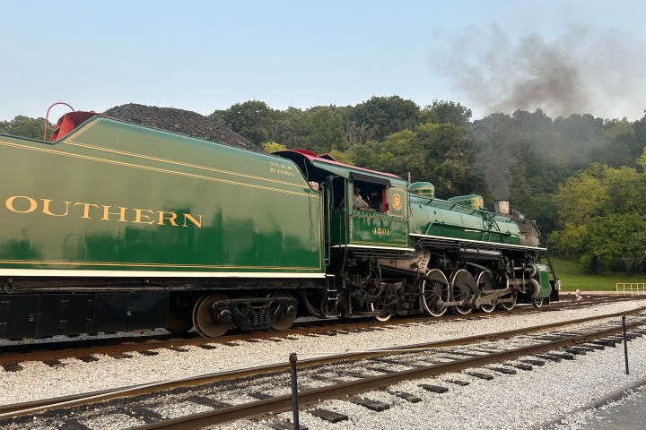 Vintage steam locomotive on tracks, emitting smoke, with trees in the background.