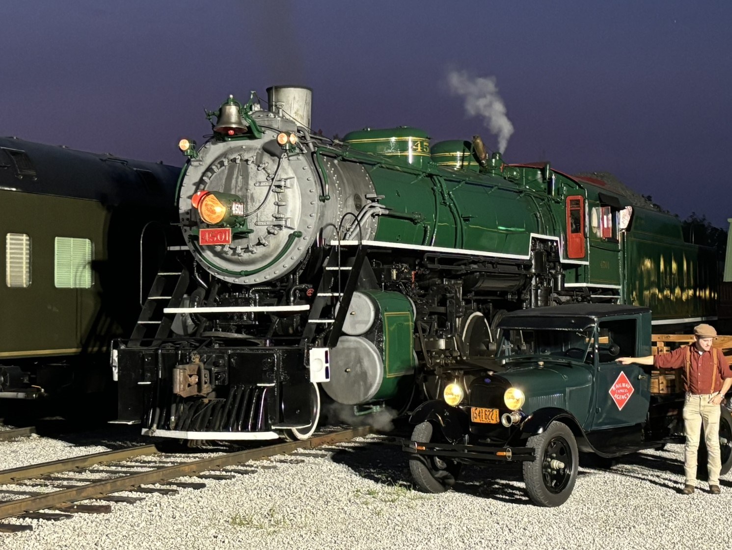 Historic steam locomotive and vintage car with man in a cap at train station.