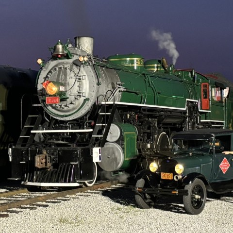 Historic steam locomotive and vintage car with man in a cap at train station.