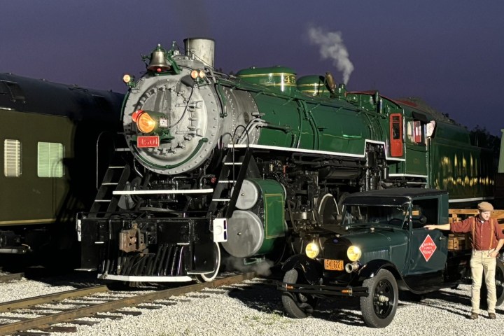 Historic steam locomotive and vintage car with man in a cap at train station.