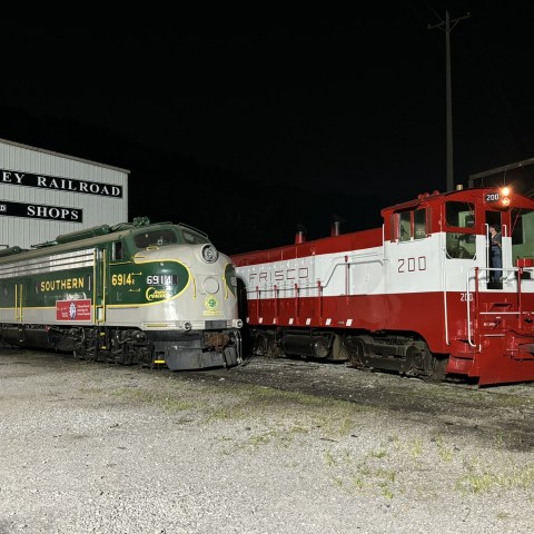 Two vintage trains parked at a rail shop, one green and one red and white, at night.