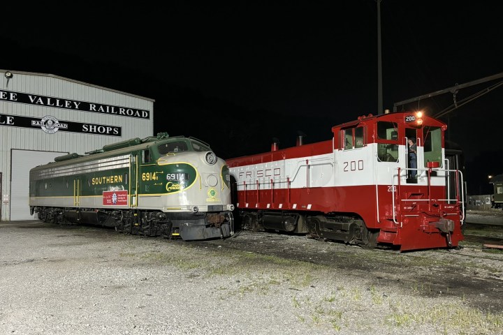 Two vintage trains parked at a rail shop, one green and one red and white, at night.