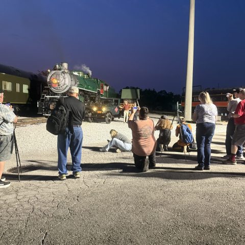 People gather near a steam locomotive at night, with a blue sky backdrop.