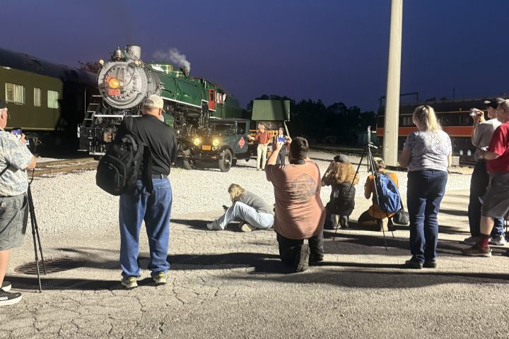 People gather near a steam locomotive at night, with a blue sky backdrop.
