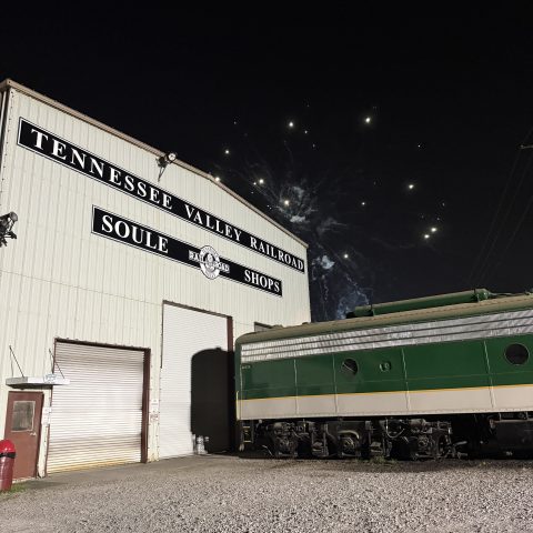Railroad shop building with a green train nearby and fireworks in the night sky.