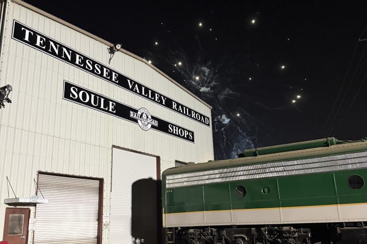Railroad shop building with a green train nearby and fireworks in the night sky.