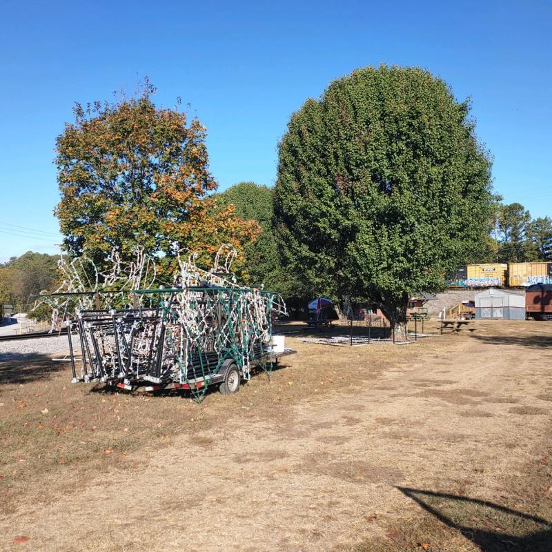 Trailer loaded with disassembled holiday light displays in an open field, with autumn trees and a freight container in the background.
