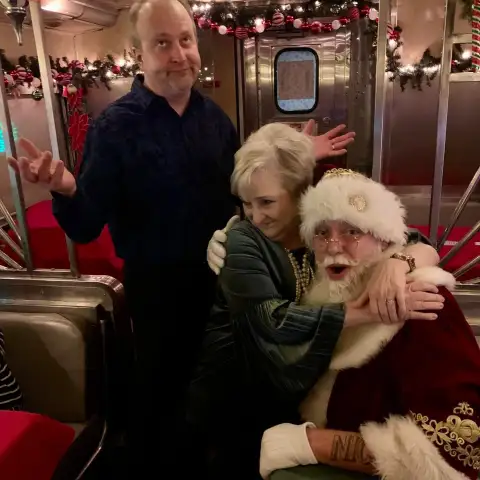 Woman hugging Santa, man shrugging in a festively decorated room.
