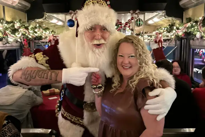 Santa and woman smiling on a festive decorated bus with lights and garlands.