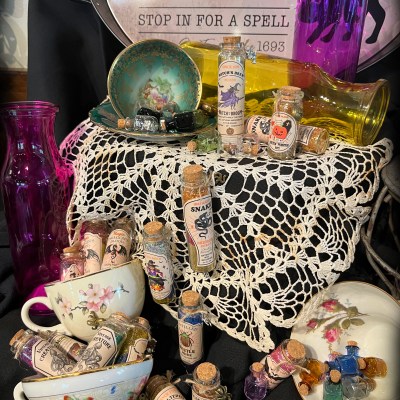 Vintage apothecary setup with potion bottles, teacups, and crochet tablecloth on black fabric.
