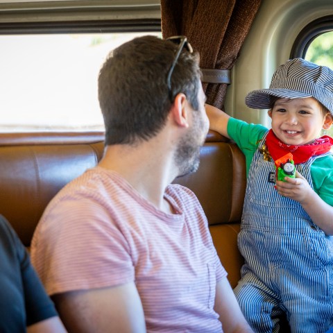 A smiling child in train conductor attire holding a toy train, sitting inside a passenger car during the Day Out with Thomas.
