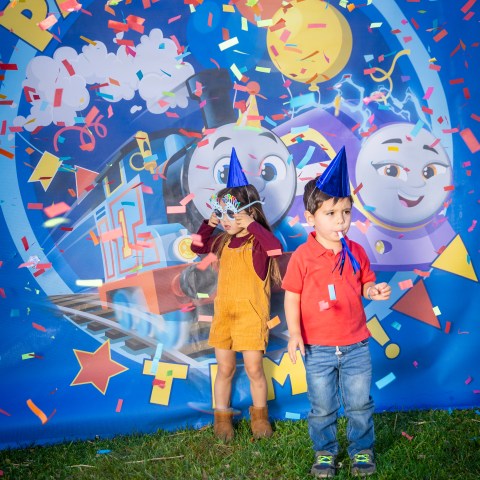 Two children standing under colorful confetti with a Thomas & Friends-themed backdrop at a party.