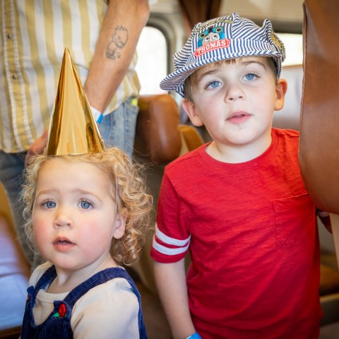 Two kids, one wearing a gold party hat and the other in a striped Thomas cap, sit on a train during the Day Out with Thomas event.