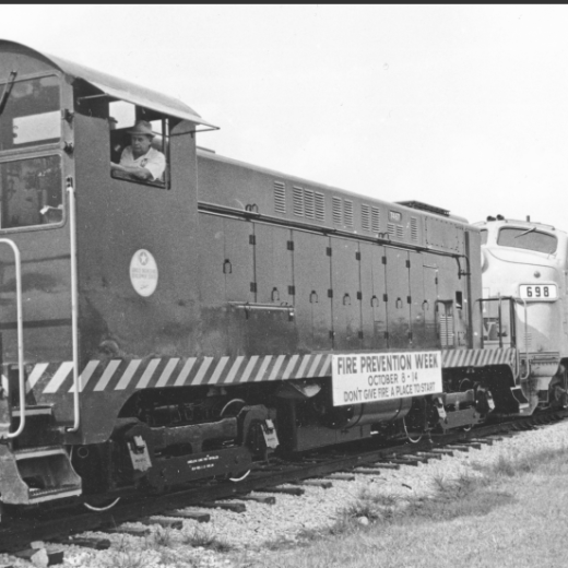 Black and white photo of a train with a 'Fire Prevention Week' sign and a conductor visible in the cab.