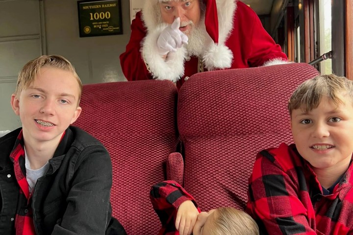 Santa playfully gestures at a train passenger with three kids in checkered shirts seated in front.