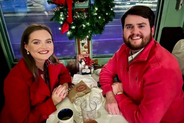 A smiling couple in red attire sits at a table with a holiday wreath on a window in the background.