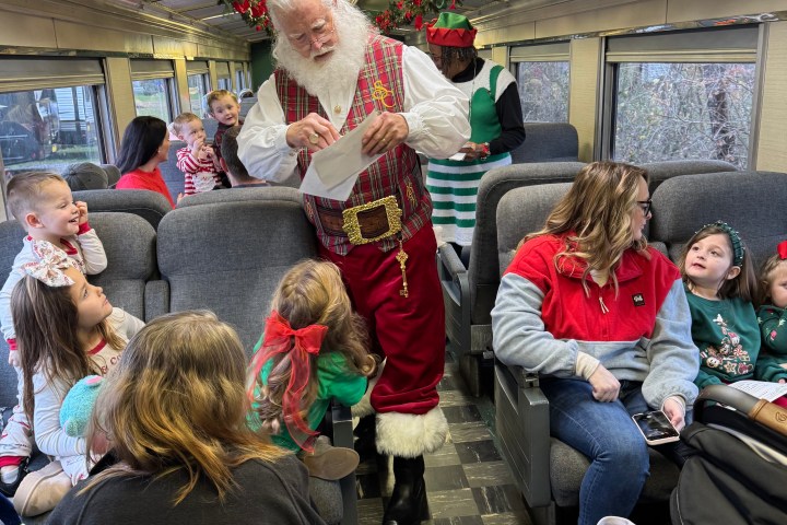 Santa interacting with children on a decorated train, with festive garlands and an elf in the background.