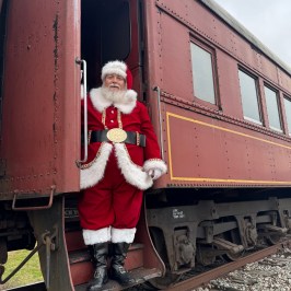 Santa Claus in a red suit greets guests at the entrance of a vintage Summerville holiday train.