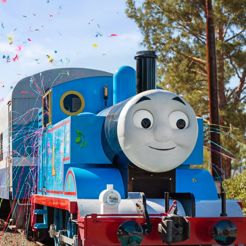 Colorful train with smiling face, surrounded by confetti and trees in the background.