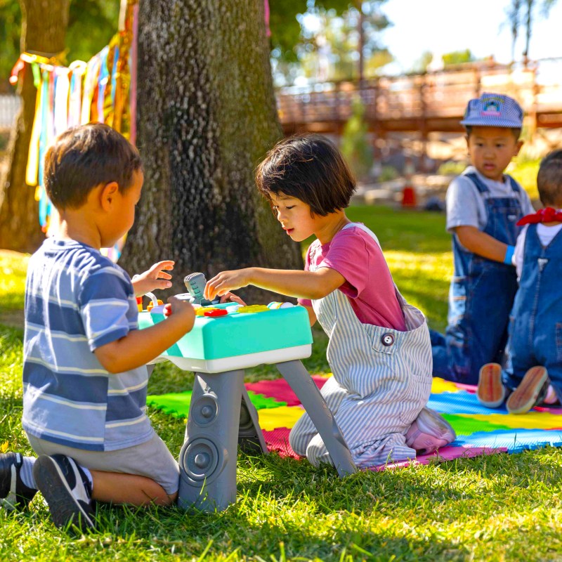 Children playing with toys on a colorful mat in a sunny park.