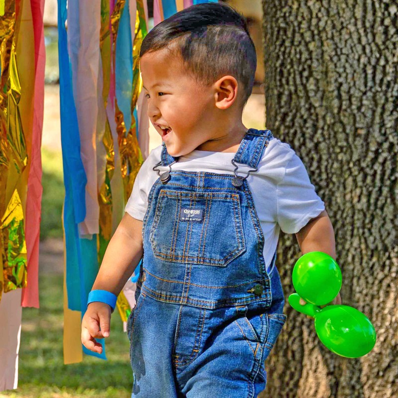 Child in denim overalls holding green balloons, near colorful streamers and tree.