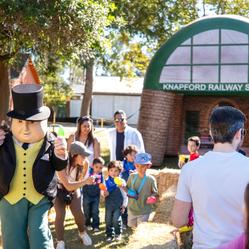 People gather around a large mascot near a Knapford Railway Station backdrop outdoors.