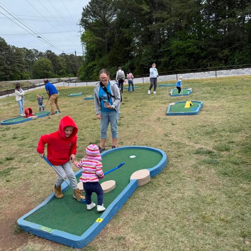 Children and adults playing mini golf in an outdoor course on a grassy field.