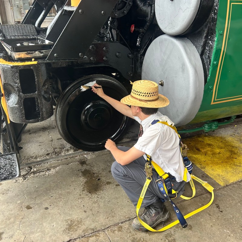 Person wearing a hat and harness paints a train wheel in a workshop.