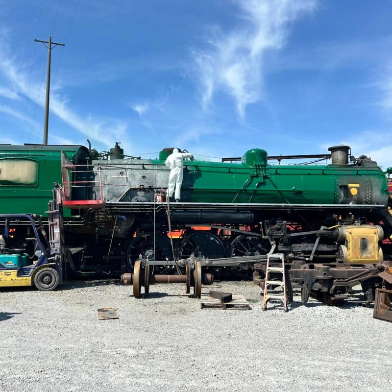 Person in white suit working on a green steam locomotive outdoors under a blue sky.