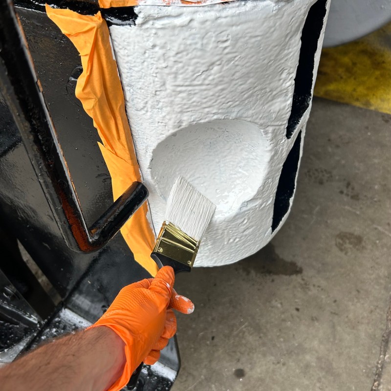 Person painting white onto a cylindrical object with a brush, wearing orange gloves.